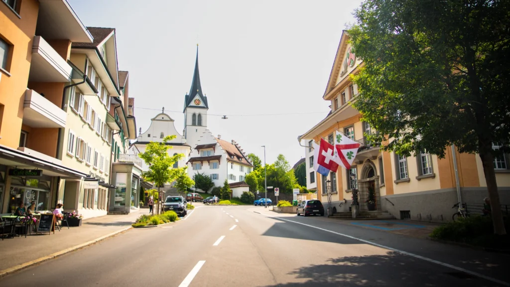 Zentrale Ansicht der Hochdorfer Hauptstrasse bei Tageslicht. Im Hintergrund erhebt sich die Pfarrkirche St. Martin mit ihrem markanten Turm. Links im Bild ist das Café Bijou mit Aussensitzplätzen zu sehen, rechts das Gemeindehaus von Hochdorf mit seiner gelben Fassade.